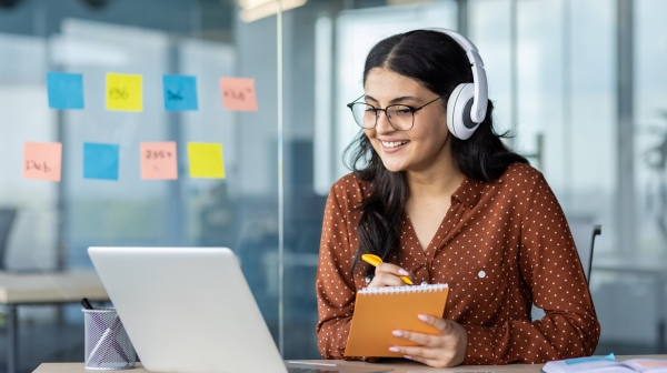 Woman watching online video course, training conference inside office at workplace. Office worker in headphones using laptop for remote meeting, writing data in notebook