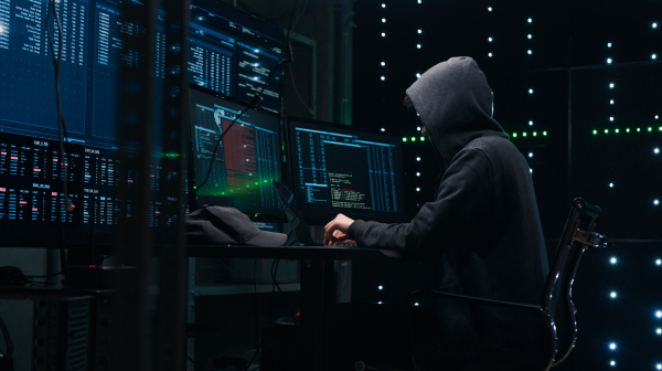 Computer hacker coding on laptop on a background of monitors.